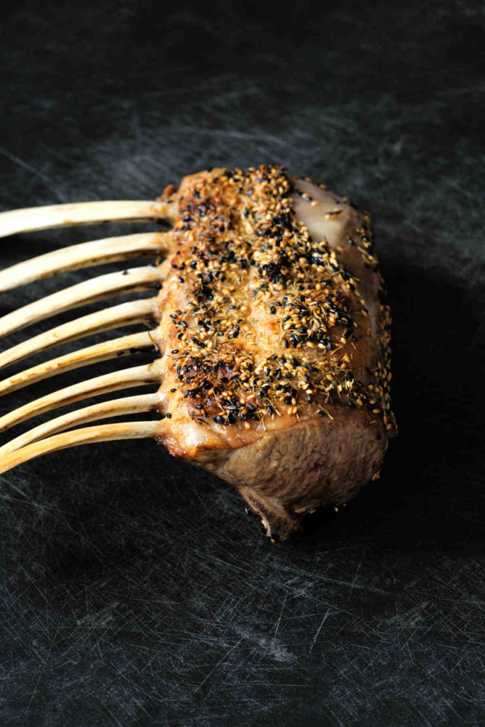 Cooked rack of lamb covered in sesame seeds on a black cutting board.