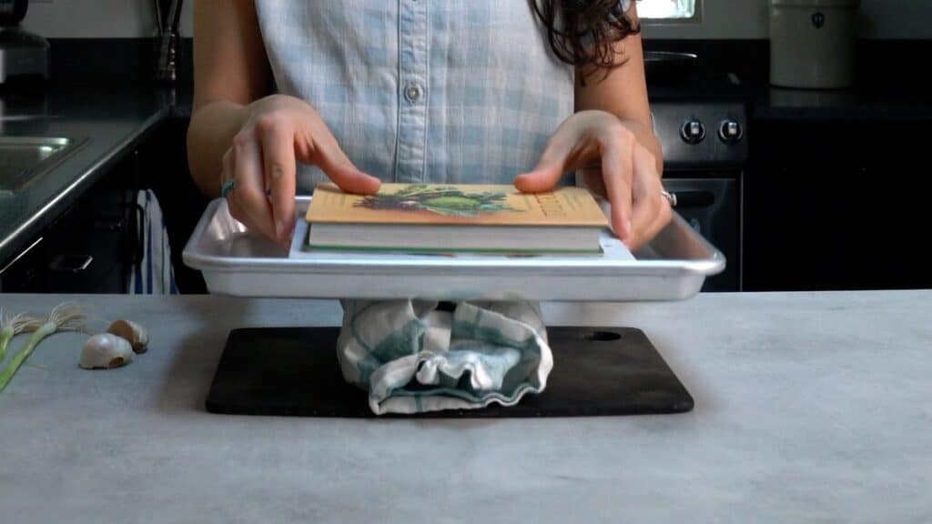 Tofu wrapped in a kitchen towel, topped with a sheet pan and books.