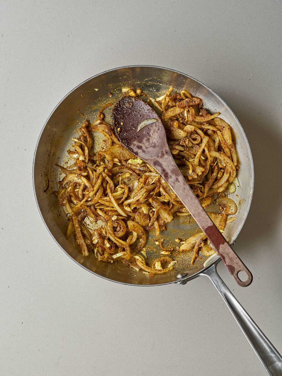 Sautéed sliced onions coated in brown and yellow shawarma spices in a metal skillet.