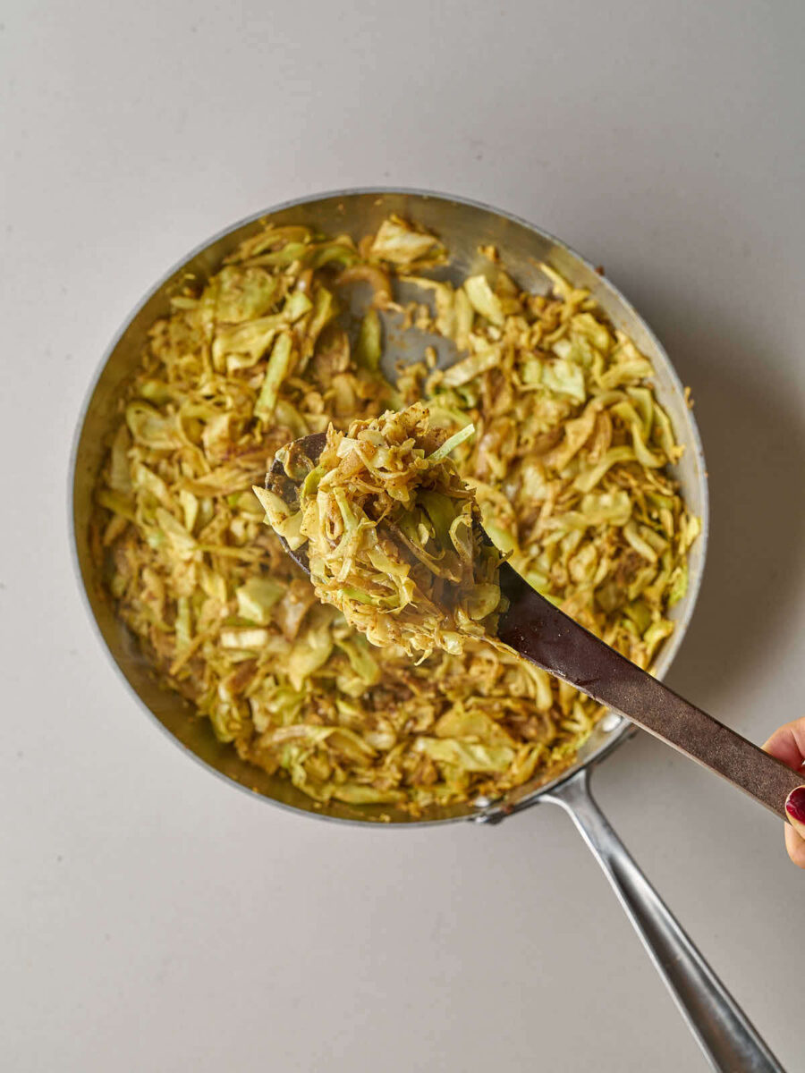 A wooden spoon lifting a serving of seasoned, sautéed shredded cabbage and onions from a skillet.