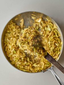 A wooden spoon holds a generous portion of seasoned, sautéed shredded cabbage and onions above the skillet they were cooked in.