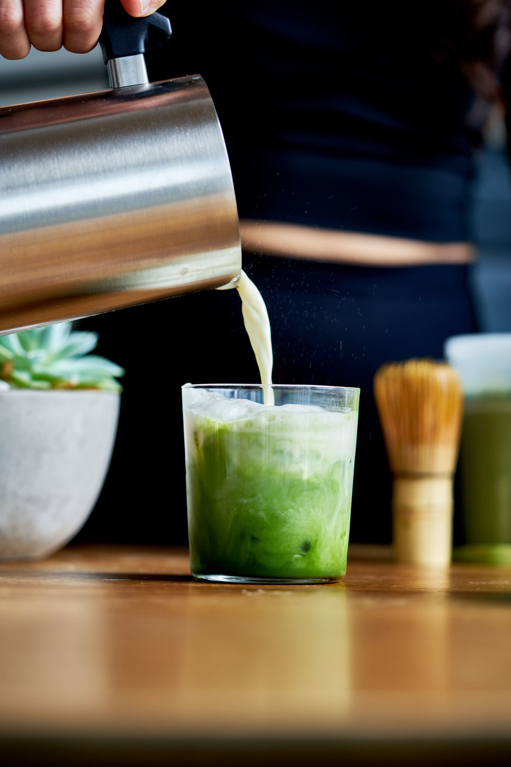 Pouring milk into an iced matcha latte on a wood table.
