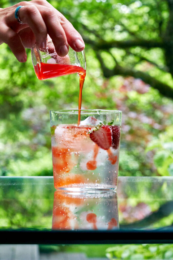 Pouring strawberry shrub over sparking water in a glass with strawberries and ice.