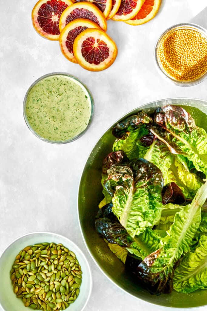 Green salad dressing next to oranges, mustard seeds, a bowl of lettuce, and pumpkin seeds.