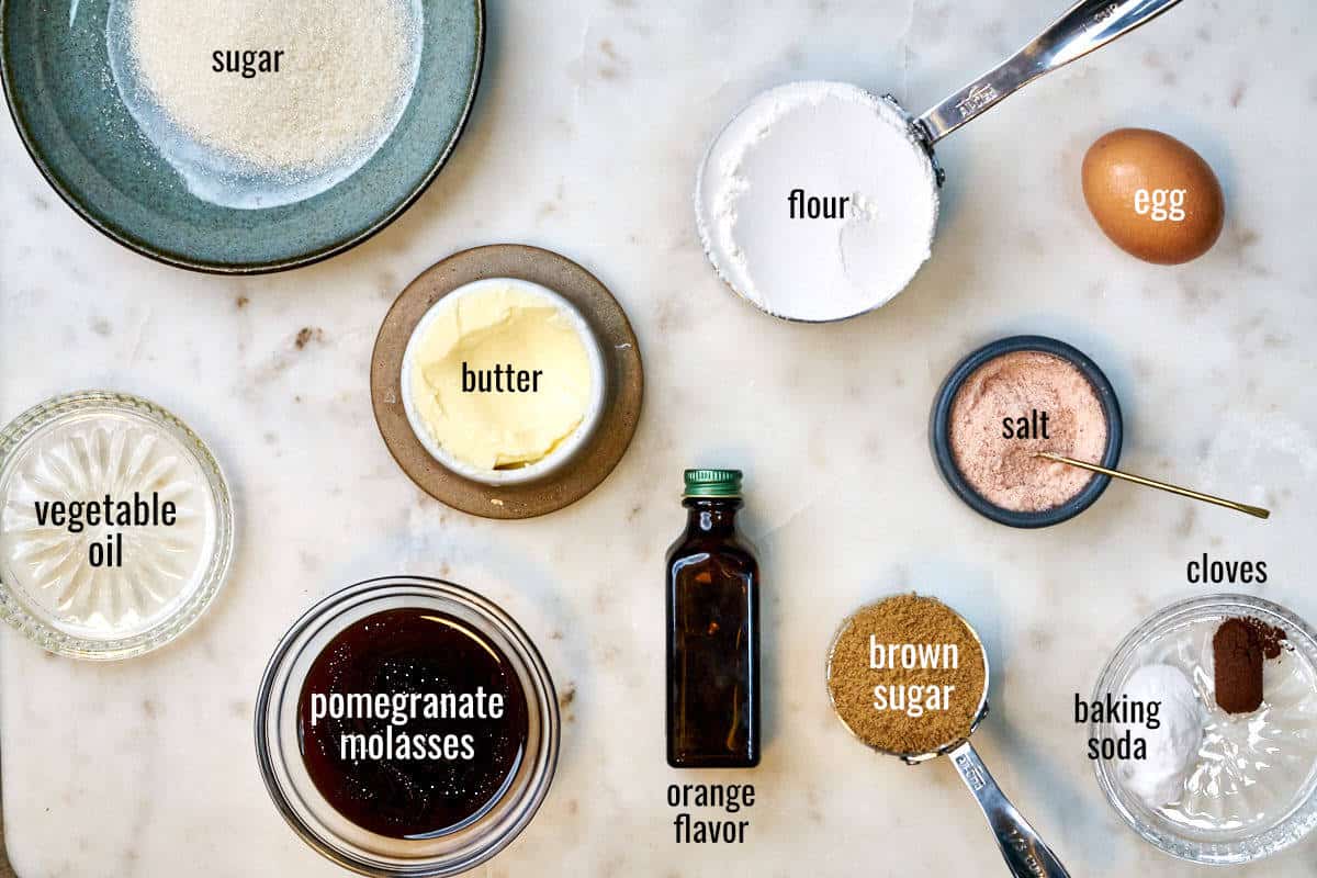 Ingredients for cookies on a white countertop including pomegranate molasses, orange flavor, and cloves.