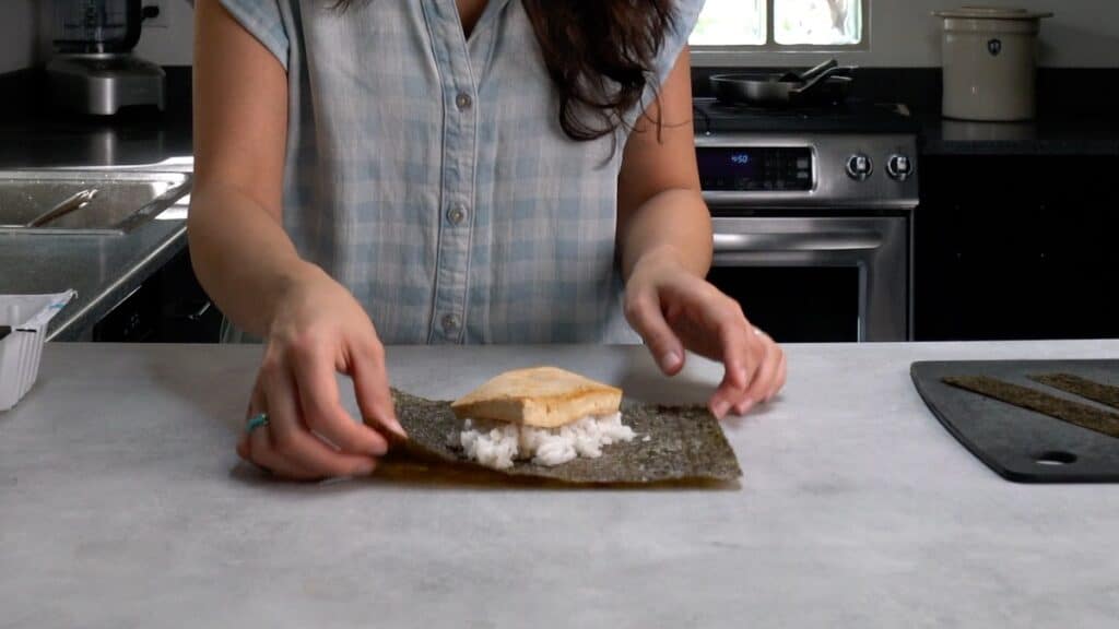 Person wrapping rice and tofu in a sheet of seaweed.