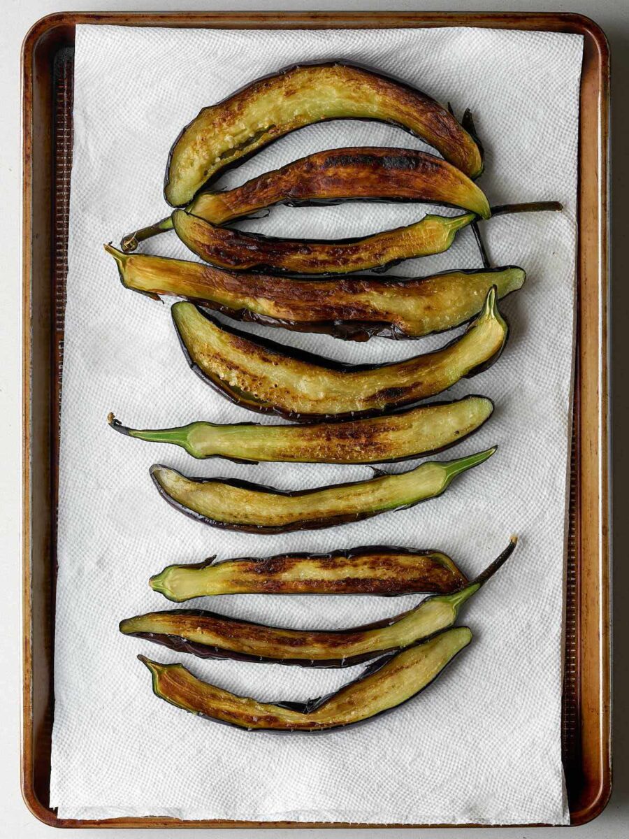 An overhead shot of a metal baking sheet lined with a paper towel, on which several long, halved pieces of cooked eggplant are arranged. The eggplant is golden brown and appears slightly charred in places.