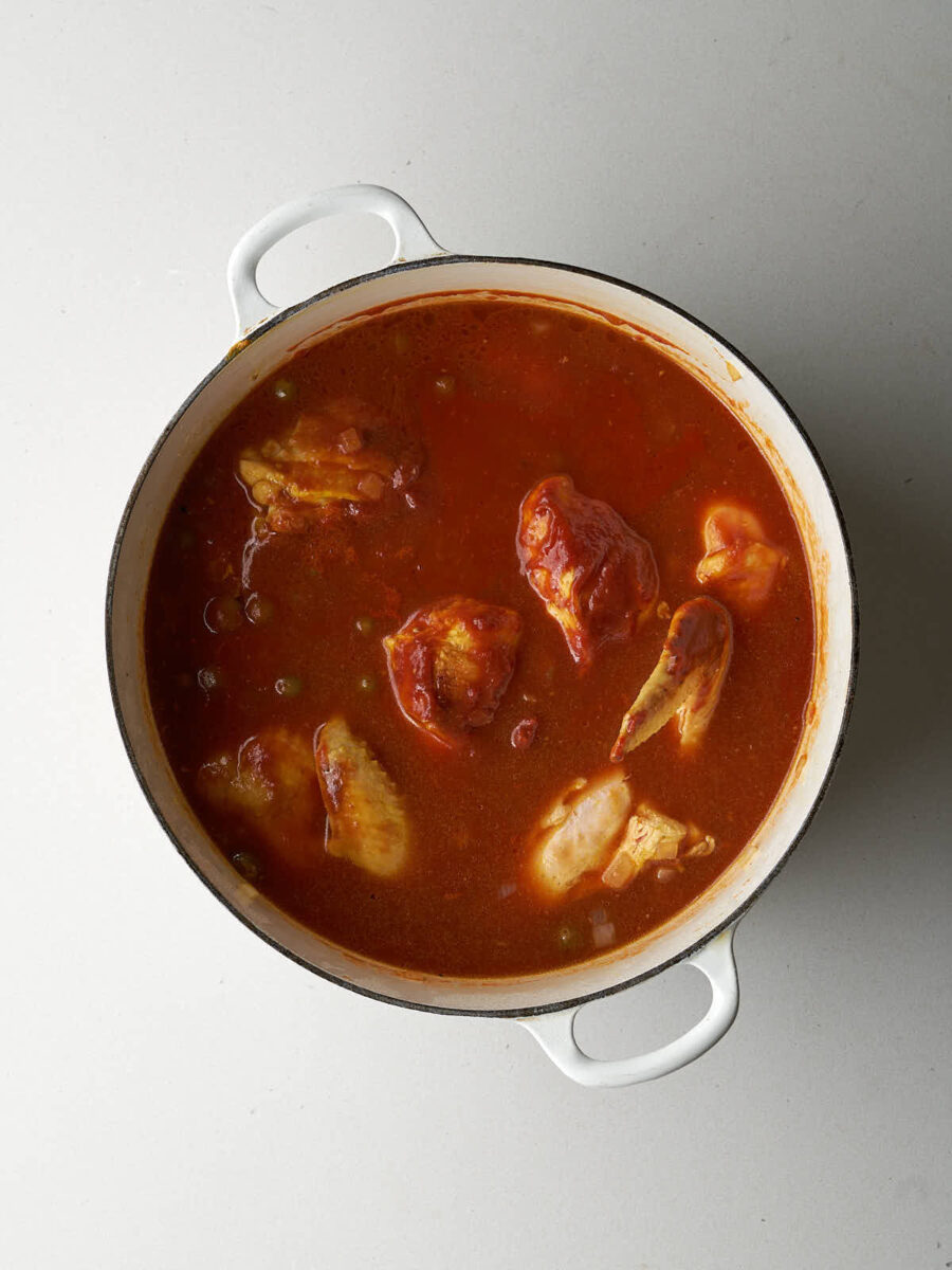 An overhead shot of a white pot filled with a vibrant reddish-orange sauce. Several pieces of chicken are submerged in the sauce, and a few green sour grames are visible. The sauce appears to be simmering.