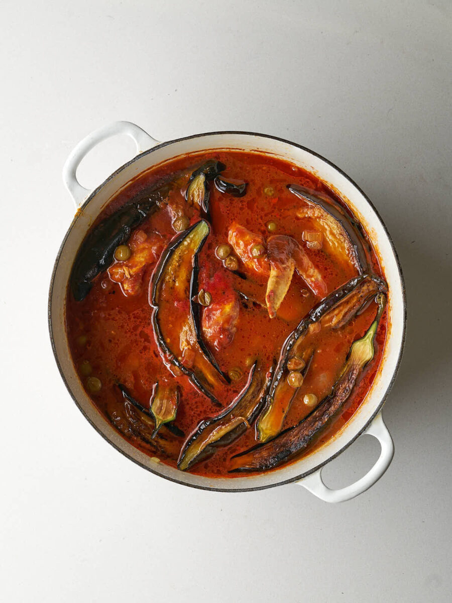 An overhead shot of a large white pot filled with a rich, reddish-orange stew. Several pieces of roasted eggplant and chicken pieces are visible in the sauce, along with some green sour grapes and eggplant.