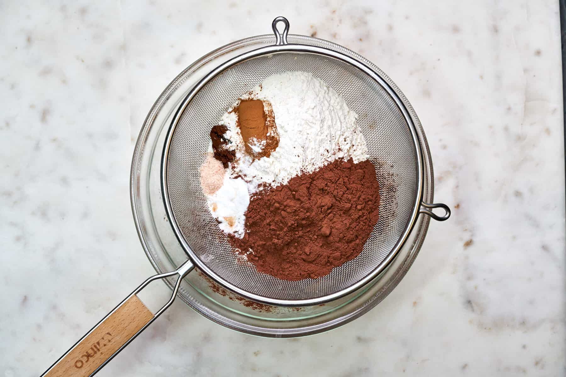 Dry cake ingredients in a strainer over a glass bowl.