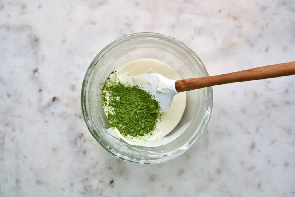 Matcha powder in a bowl with melted white chocolate and a rubber spatula.