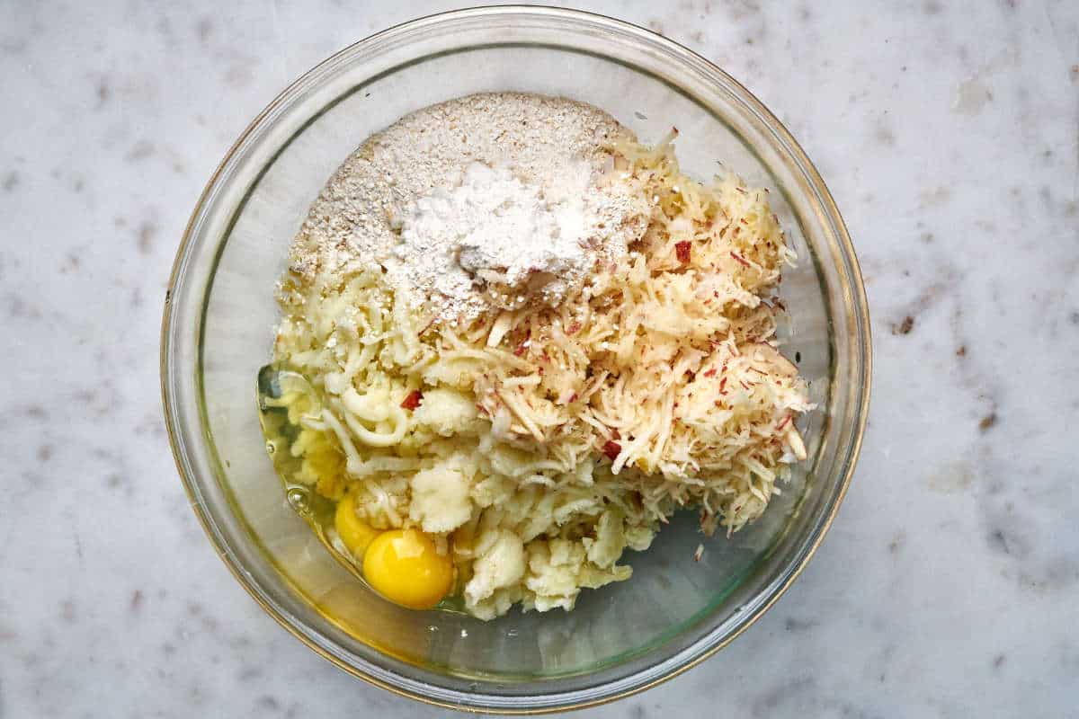 Mashed potatoes, grated apple, matzo meal, and two eggs in a glass bowl on a white countertop.