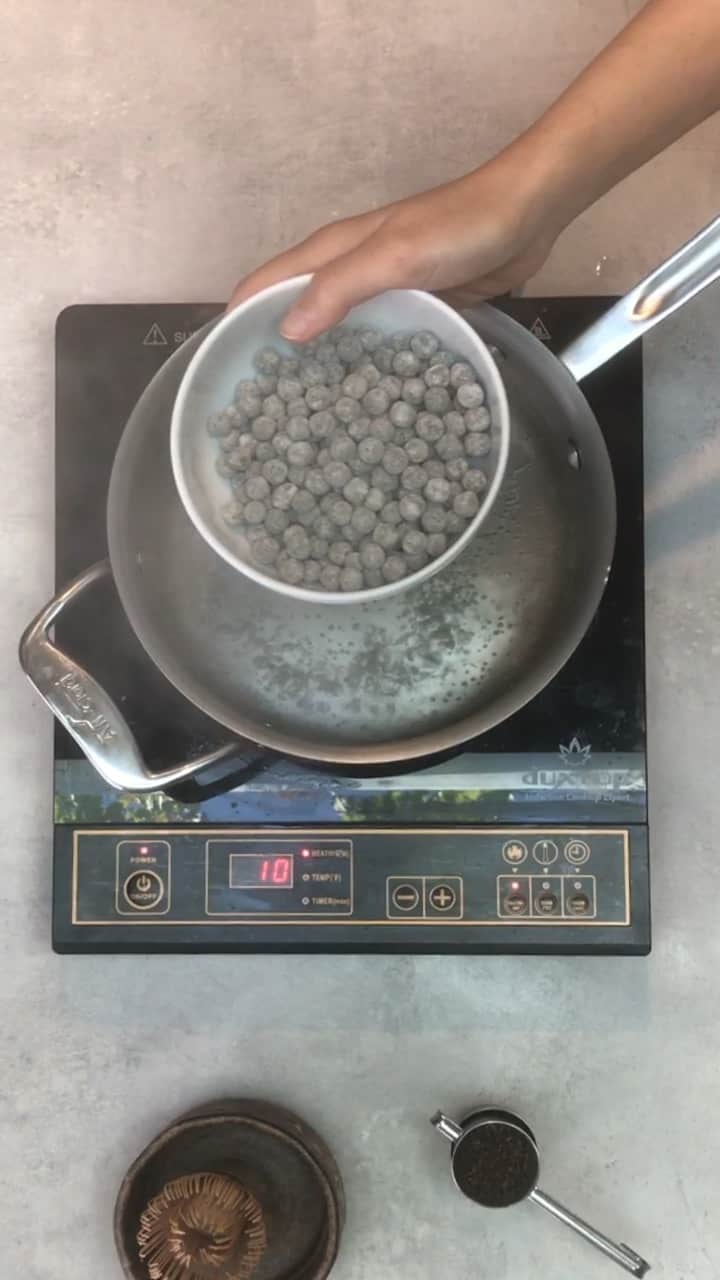 A hand holding a white bowl of tapioca pearls over a saucepan of boiling water.