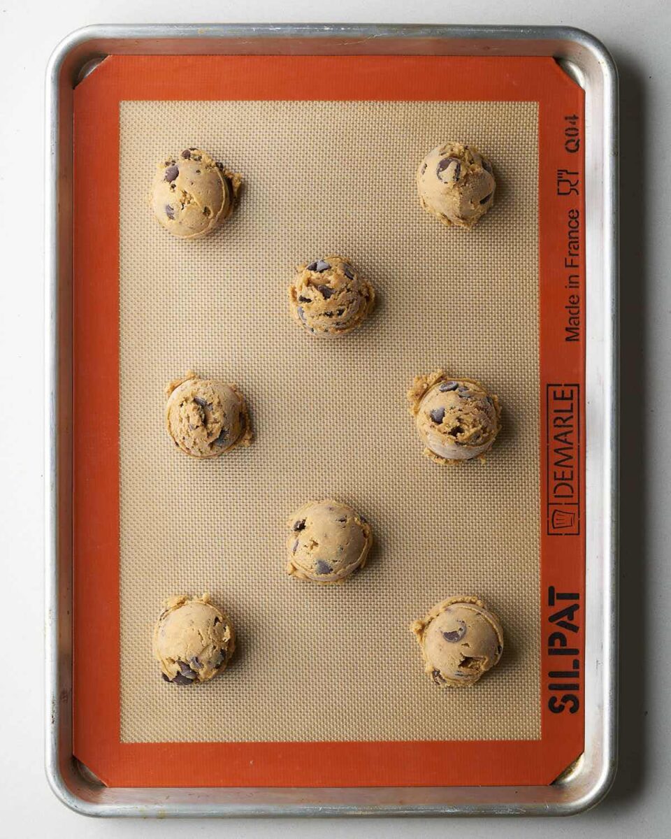 Eight scoops of cookie dough with dark chocolate chips spaced out on a baking sheet lined with a beige silicone baking mat.