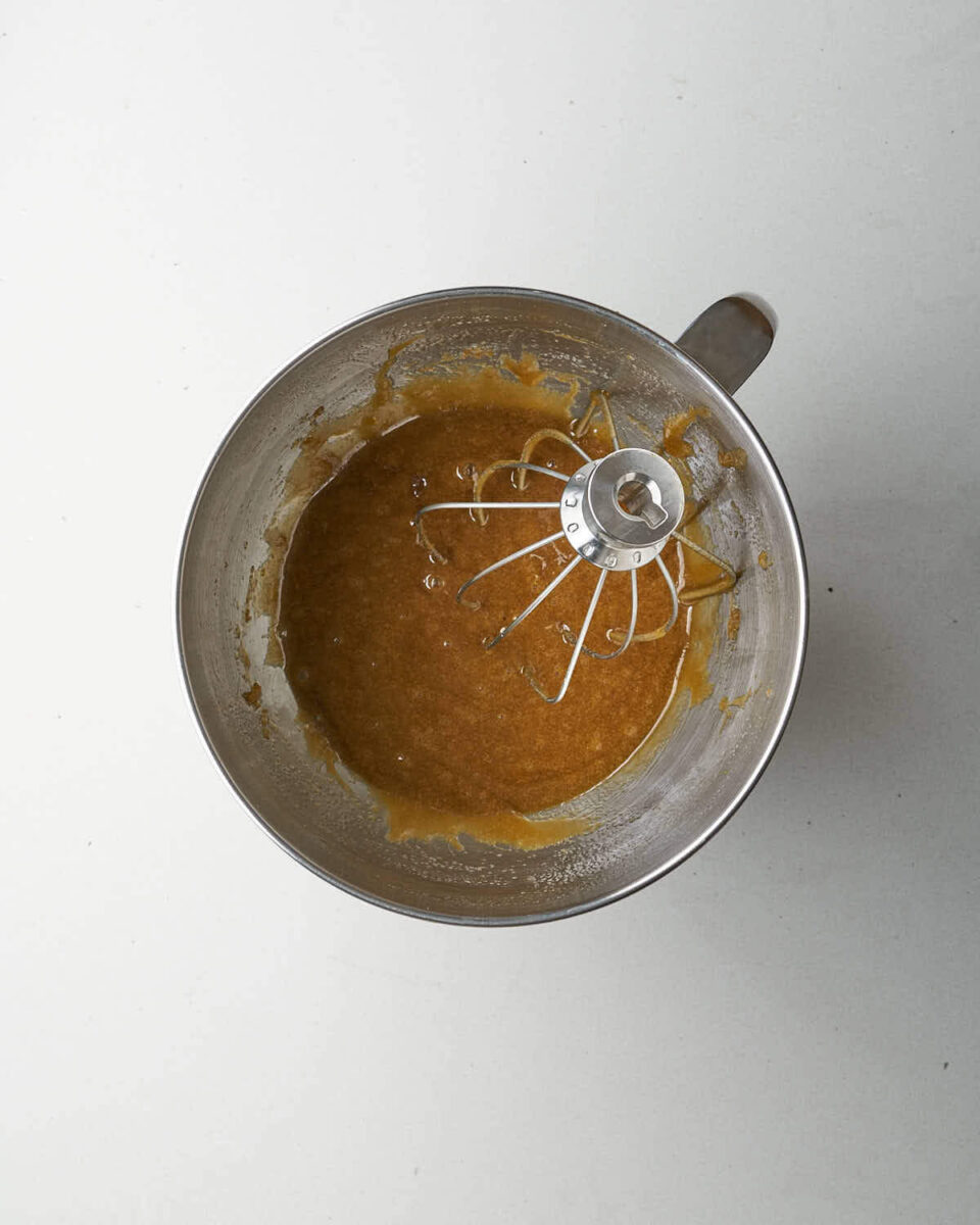 A metal mixing bowl with a whisk attachment, containing whisked wet ingredients and sugar.