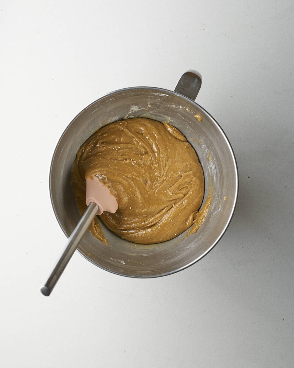 A metal mixing bowl containing smooth, light brown cookie batter with a pink spatula resting in it.