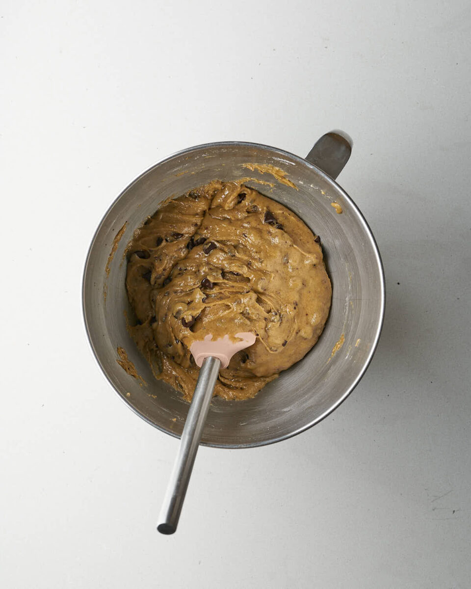 A metal mixing bowl containing finished dark chocolate chip cookie dough with a pink spatula resting in it.