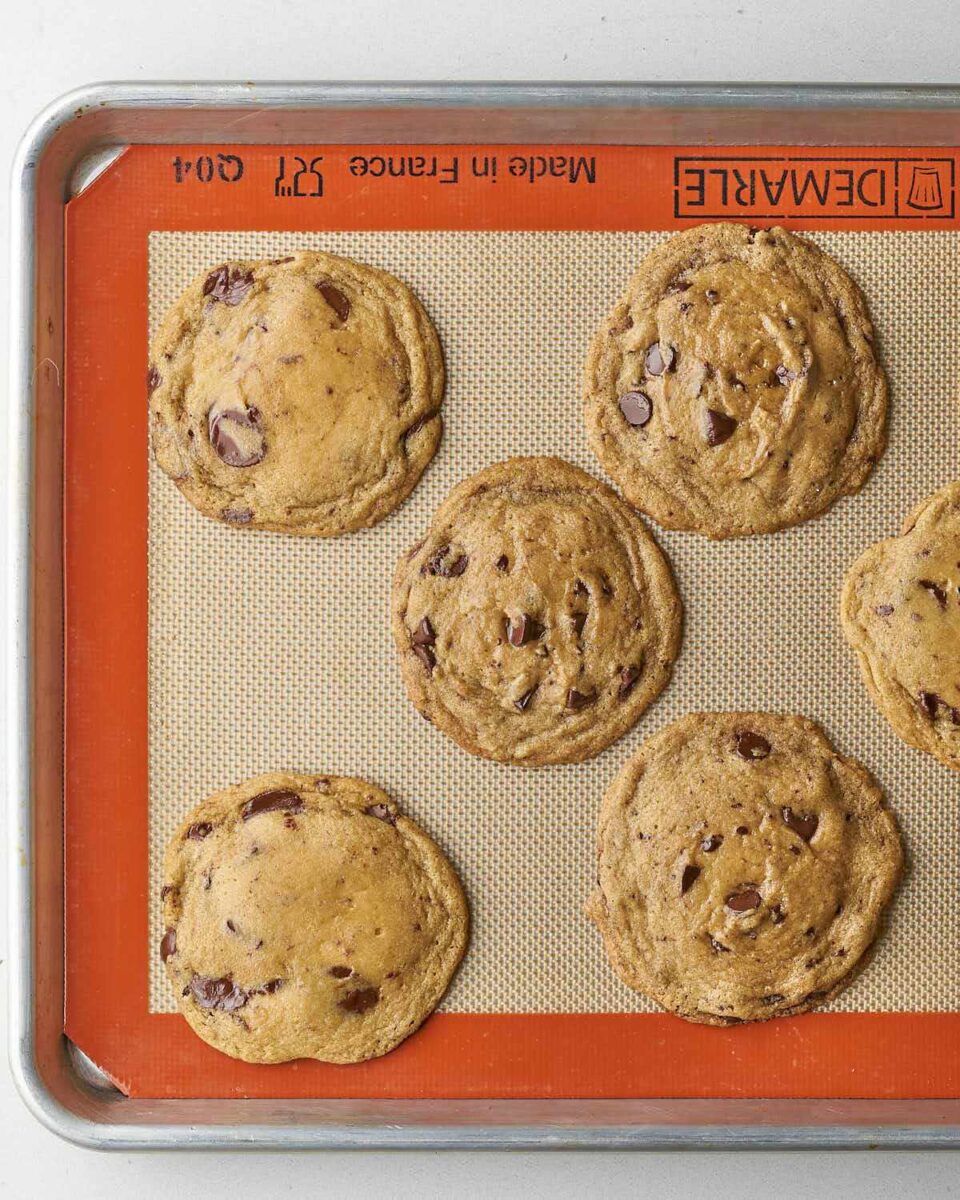 A close-up overhead photo of five dark chocolate chip cookies baked on an orange silicone mat on a metal baking sheet.