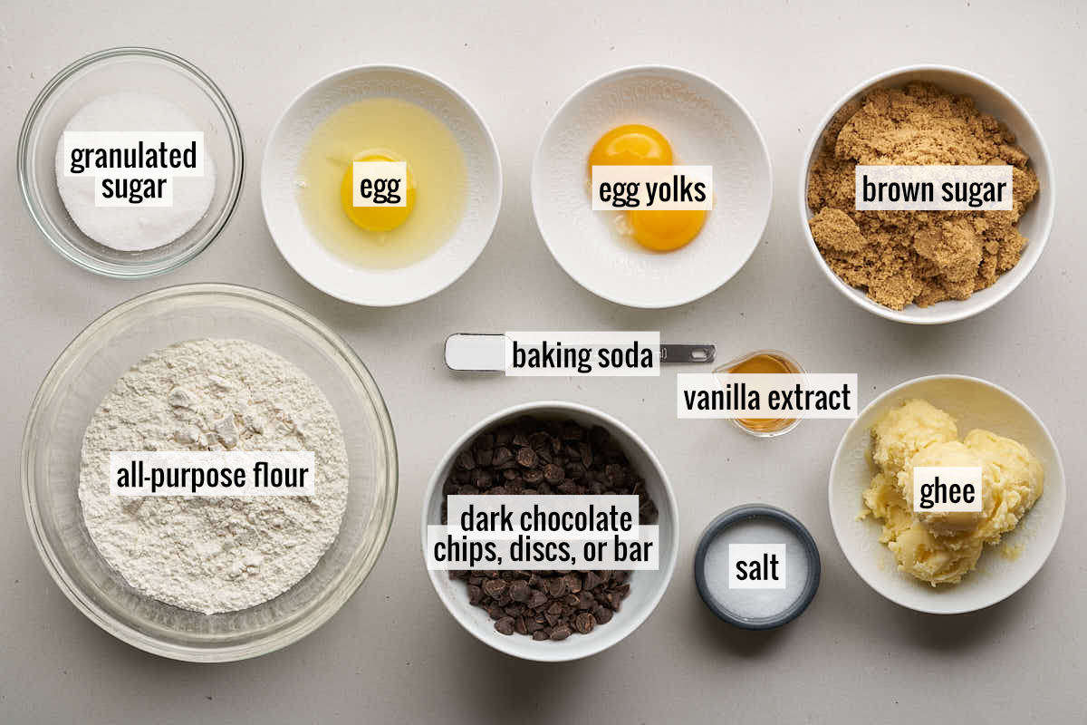 Various ingredients for dark chocolate chip ghee cookies, labeled with text. Ingredients include granulated sugar, egg, egg yolks, brown sugar, all-purpose flour, dark chocolate chips, baking soda, vanilla extract, salt, and ghee.