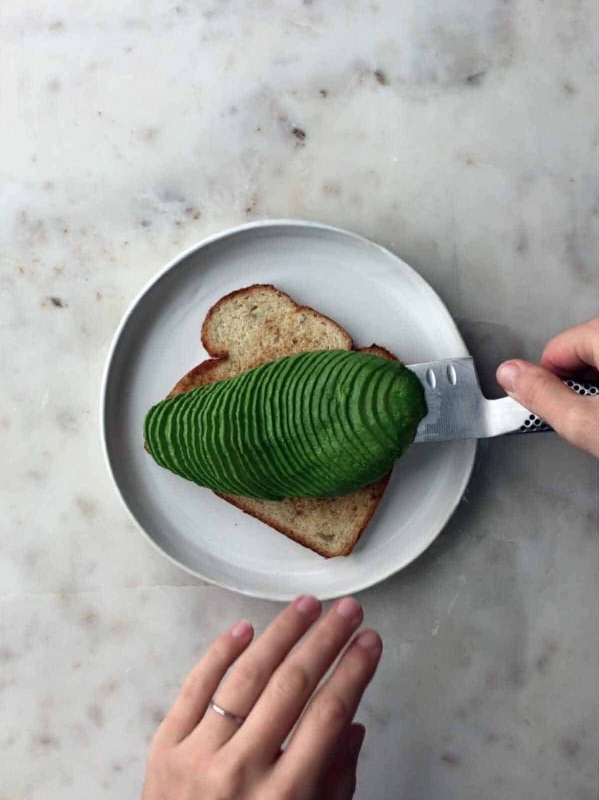 Sliced avocado being placed on a slice of toast by a hand holding it on a knife.