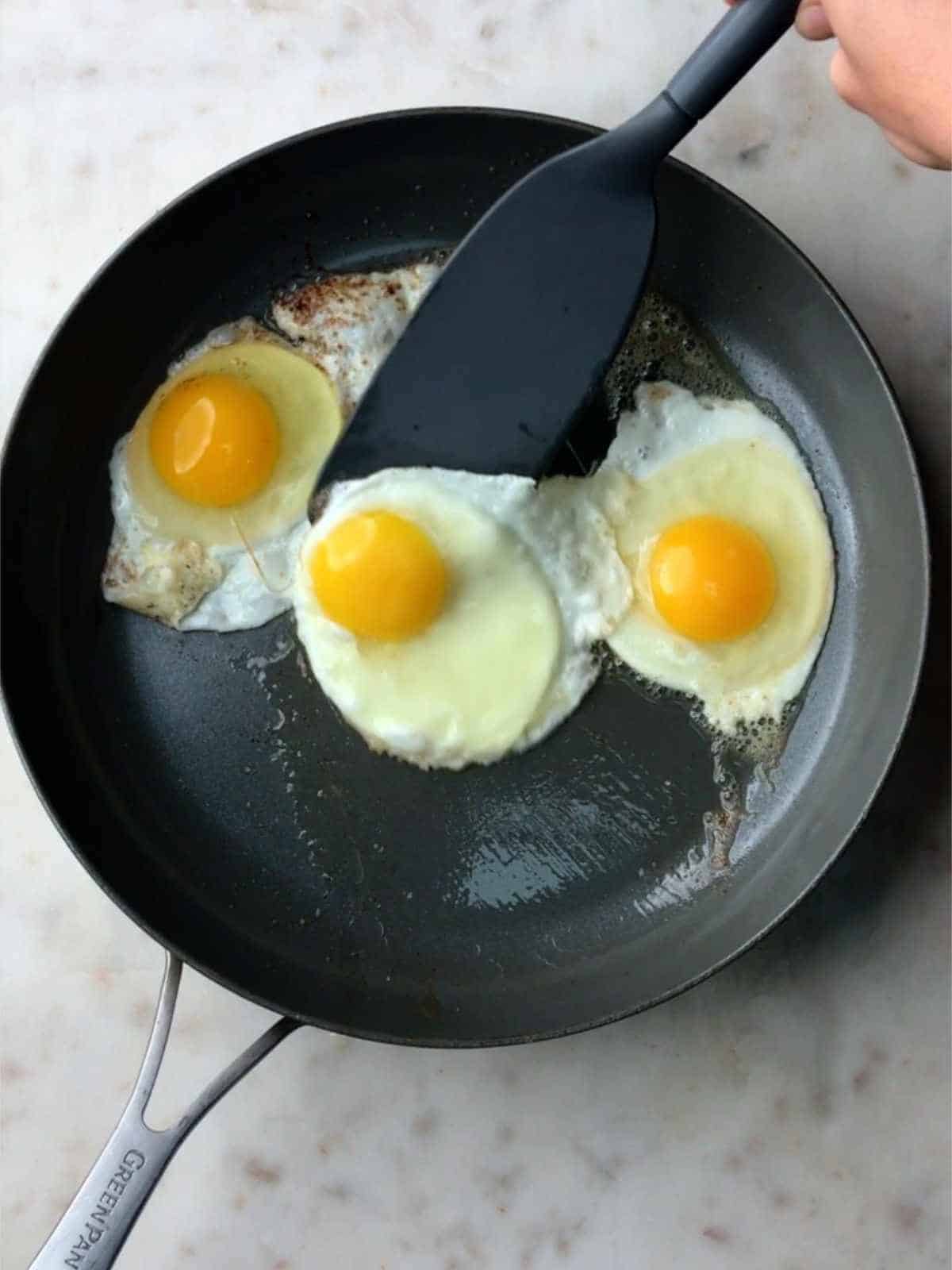 Three fried eggs in a non-stick pan with a spatula under the one in the middle.