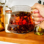 Brewed tea in a glass teapot above a wood floating shelf.