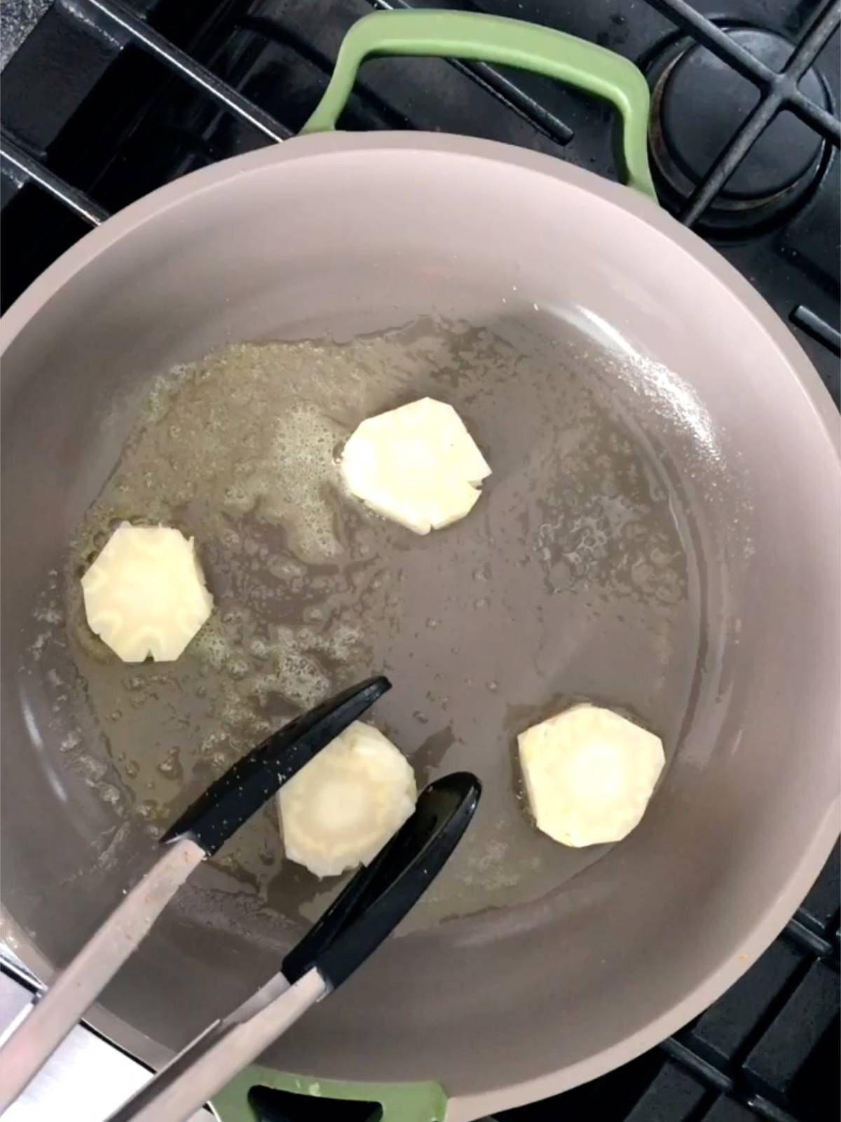 Tongs flipping a disc of cauliflower stalk in a non-stick pan with 3 other discs of cauliflower.