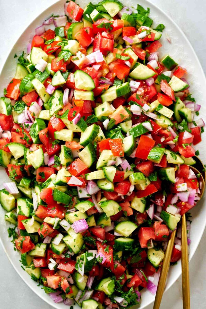 Large platter of cucumber tomato salad with gold utensils.