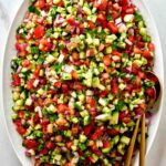 Large platter of cucumber tomato salad with gold utensils.