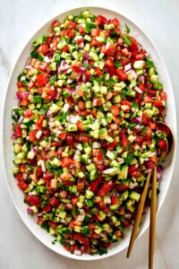 Large platter of cucumber tomato salad with gold utensils.