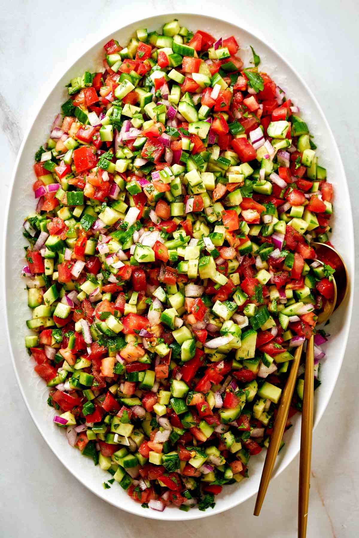 Large platter of cucumber tomato salad with gold utensils.