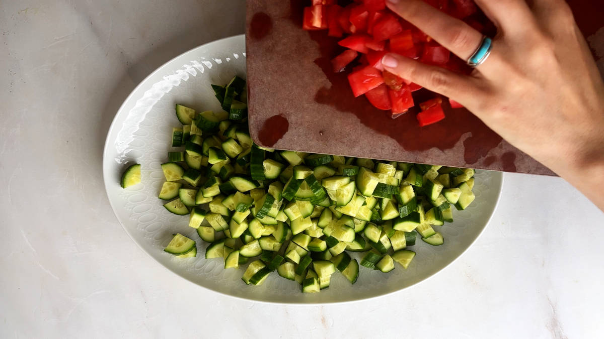 Adding diced tomatoes to a plate of diced cucumbers.
