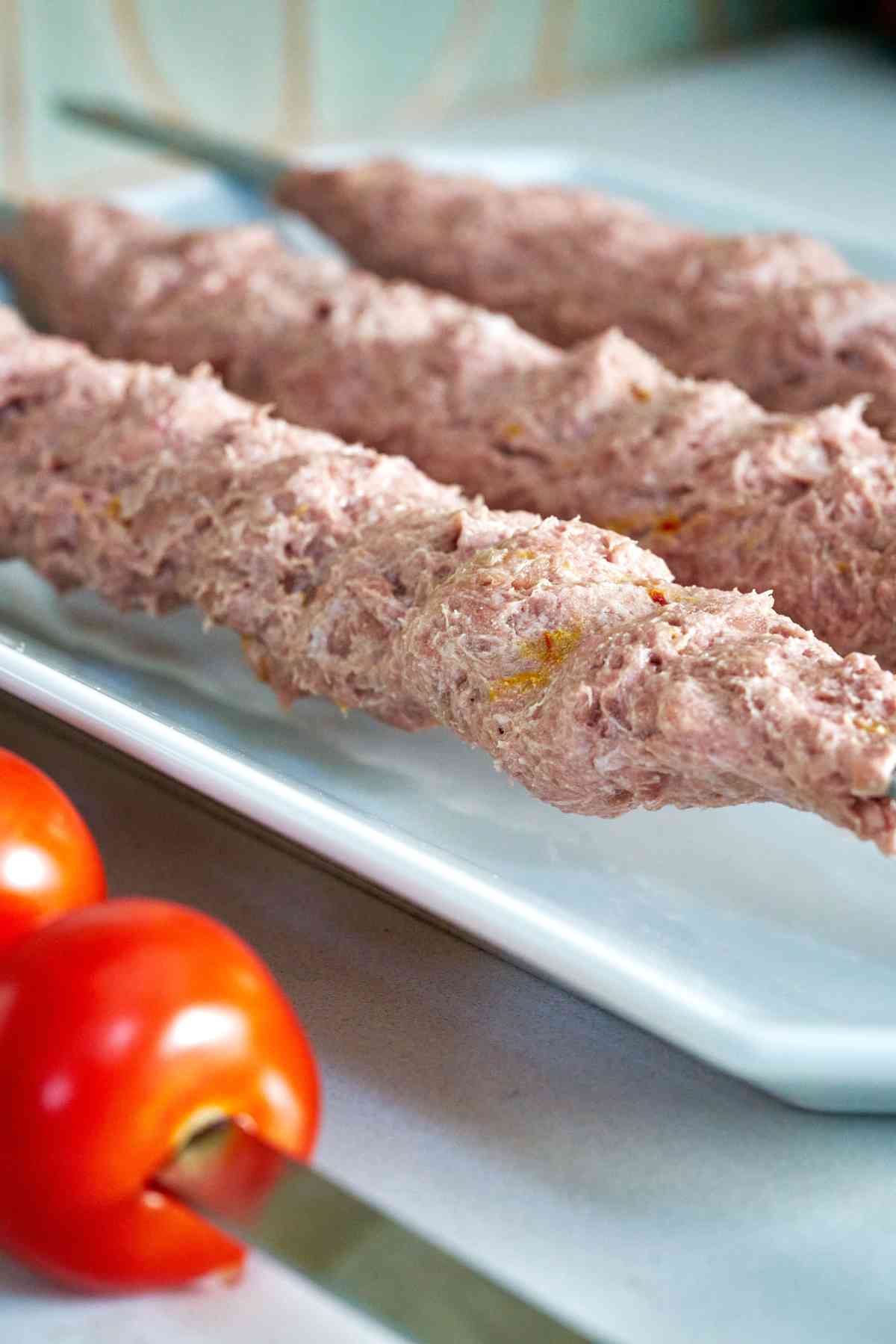 Raw meat on skewers on a plate next to tomatoes.