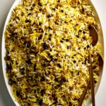 Yellow rice with lentils and currants in serving dish with gold spoons.