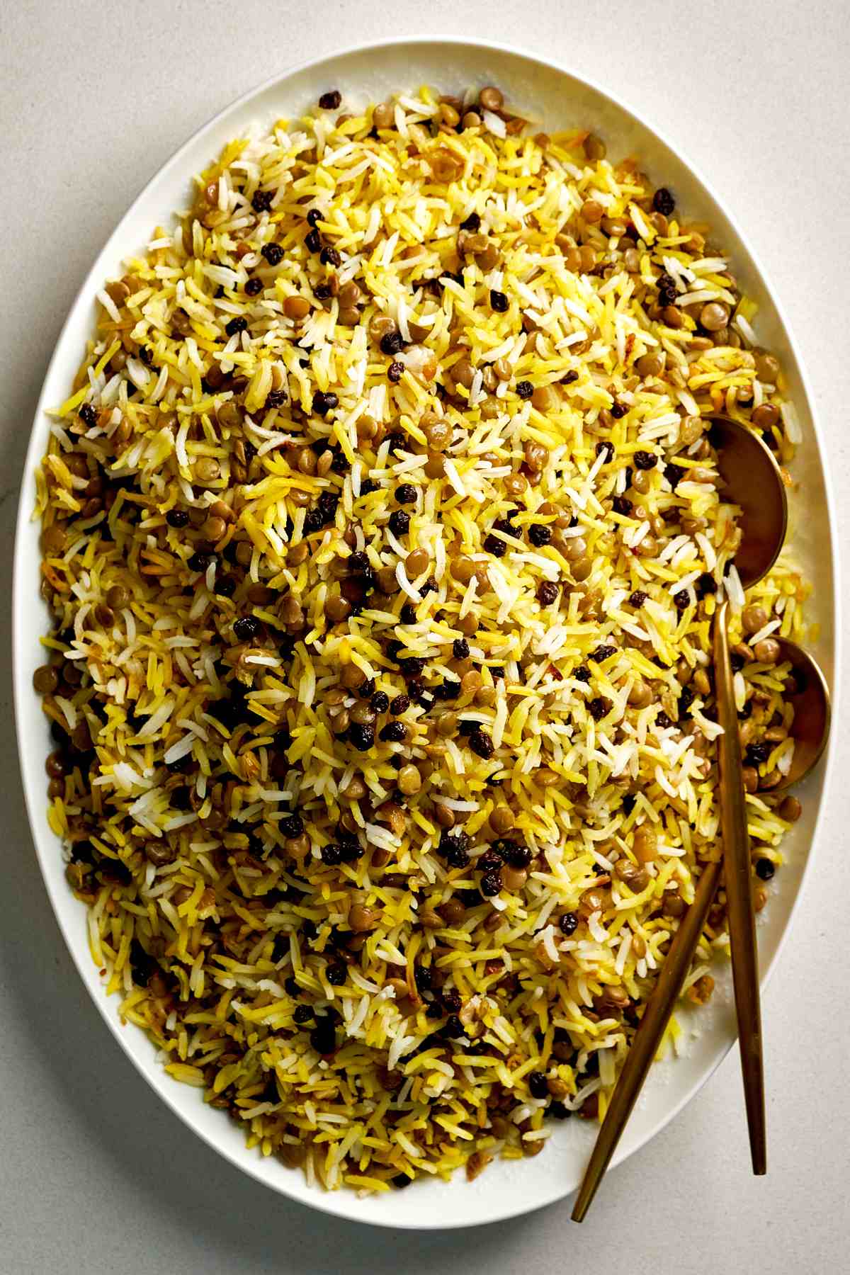 Yellow rice with lentils and currants in serving dish with gold spoons.