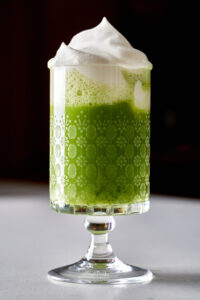 Spiked matcha cocktail with whipped cream in a pedestal glass.