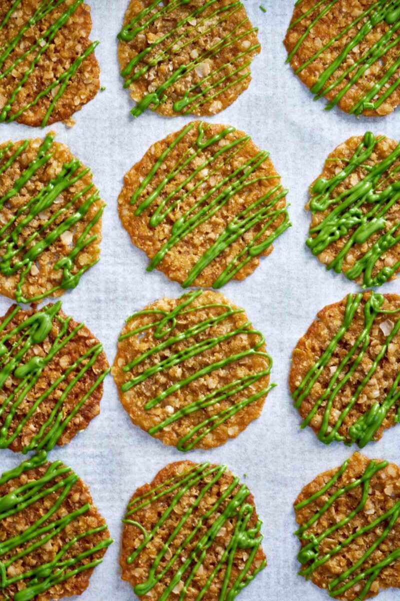 Lace cookies drizzled with green chocolate on a baking sheet.