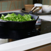 Black induction cooktop with cast iron pan filled with greens.