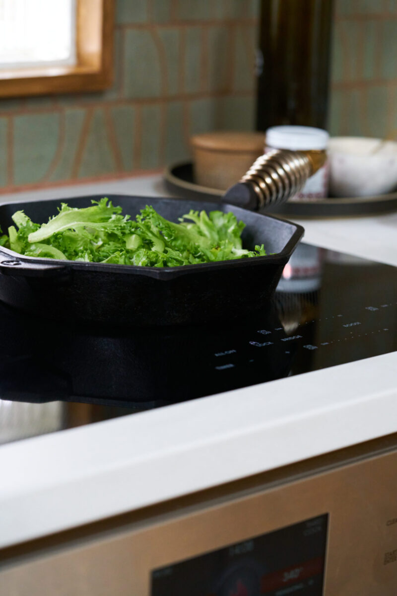 Black induction cooktop with cast iron pan filled with greens.