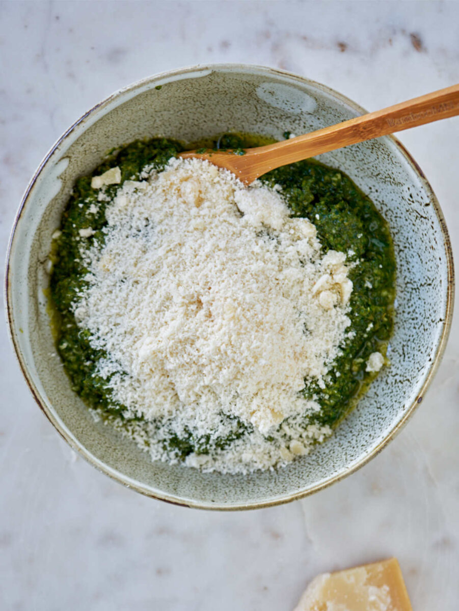 Green pesto in a ceramic bowl on a white countertop next to a parmesan wedge.