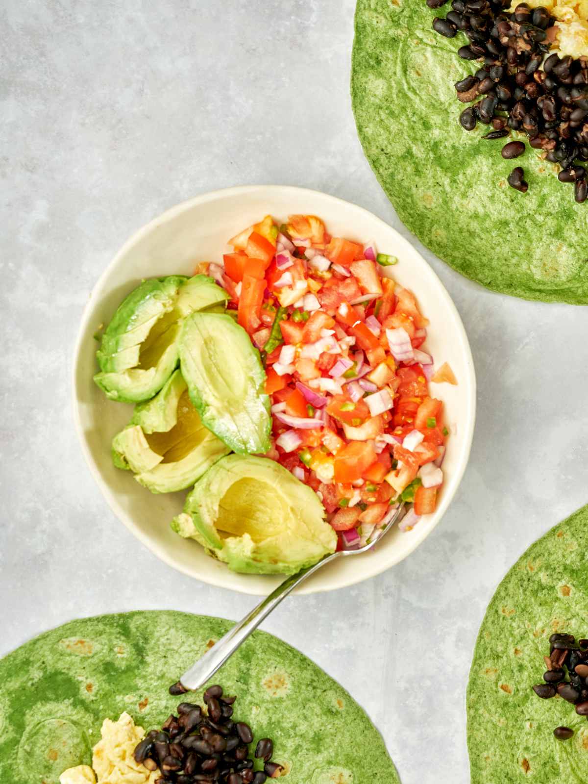 Bowl of salsa and avocado next to tortillas topped with black beans and eggs.