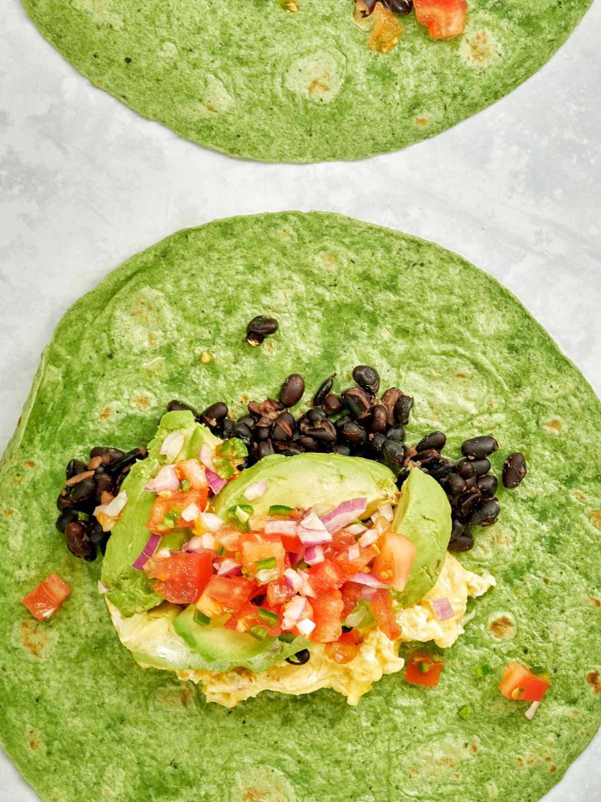 Green tortilla topped with scrabled eggs on the bottom, then avocado and black beans, and salsa on top.