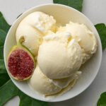 An overhead shot of a white bowl filled with four scoops of pale yellow ice cream and half a fig. A fig leaf is placed under the bowl to the left and another to the right.