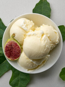 An overhead shot of a white bowl filled with four scoops of pale yellow ice cream and half a fig. A fig leaf is placed under the bowl to the left and another to the right.