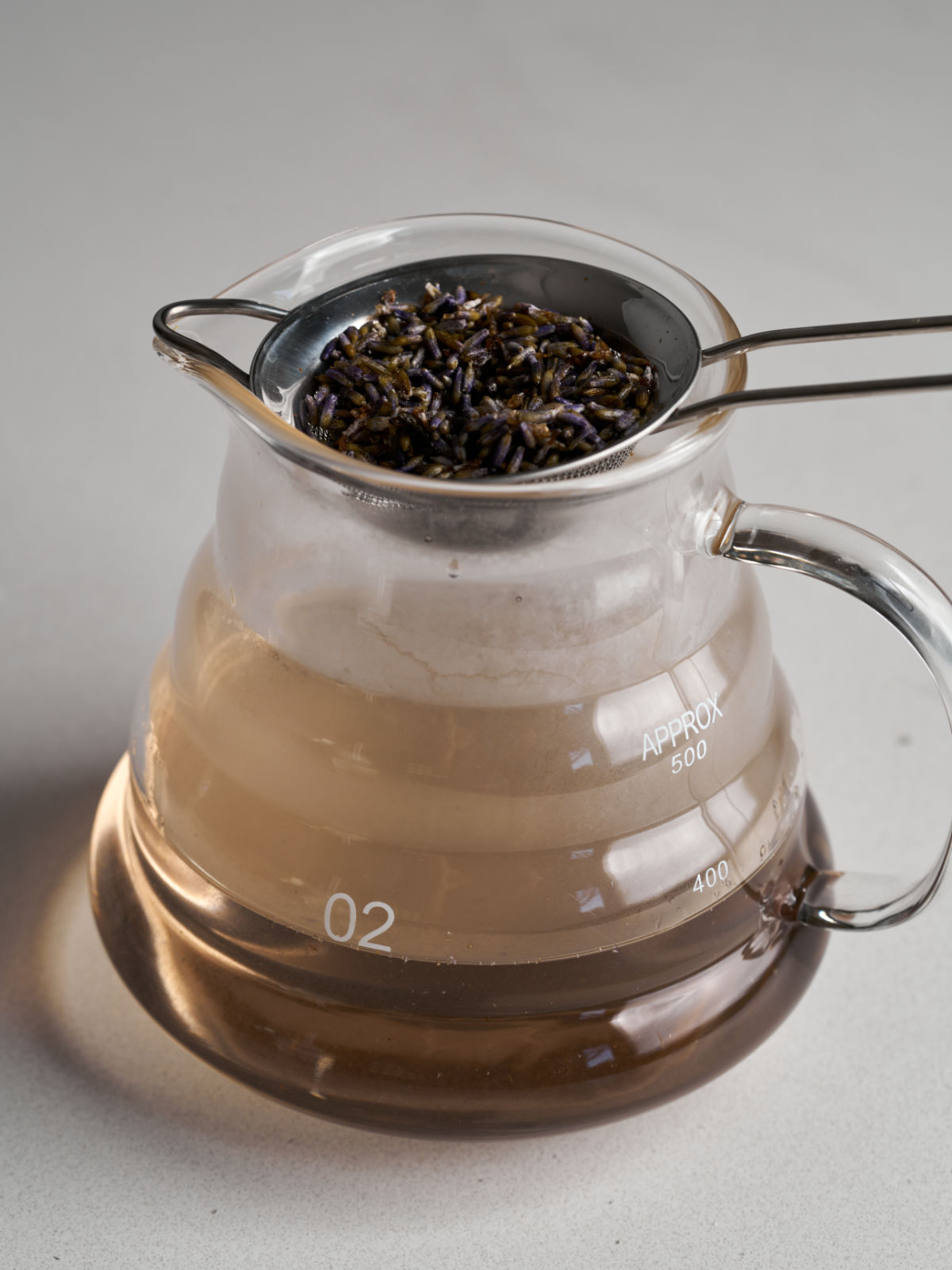 Small glass pitcher filled with brown simple syrup with a strainer on top filled with lavender flowers.