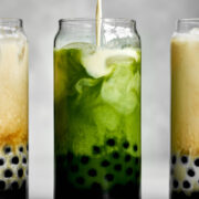 Pouring milk into a green milk tea with tapioca pearls next to two milk teas.