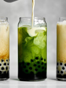 Pouring milk into a green milk tea with tapioca pearls next to two milk teas.