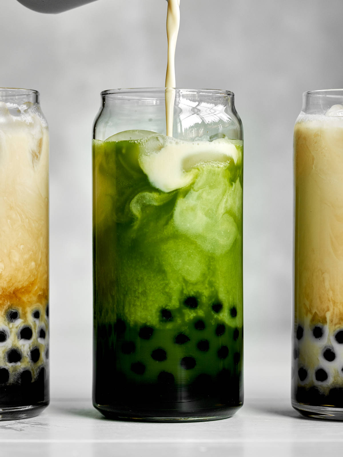 Pouring milk into a green milk tea with tapioca pearls in between two milk teas.