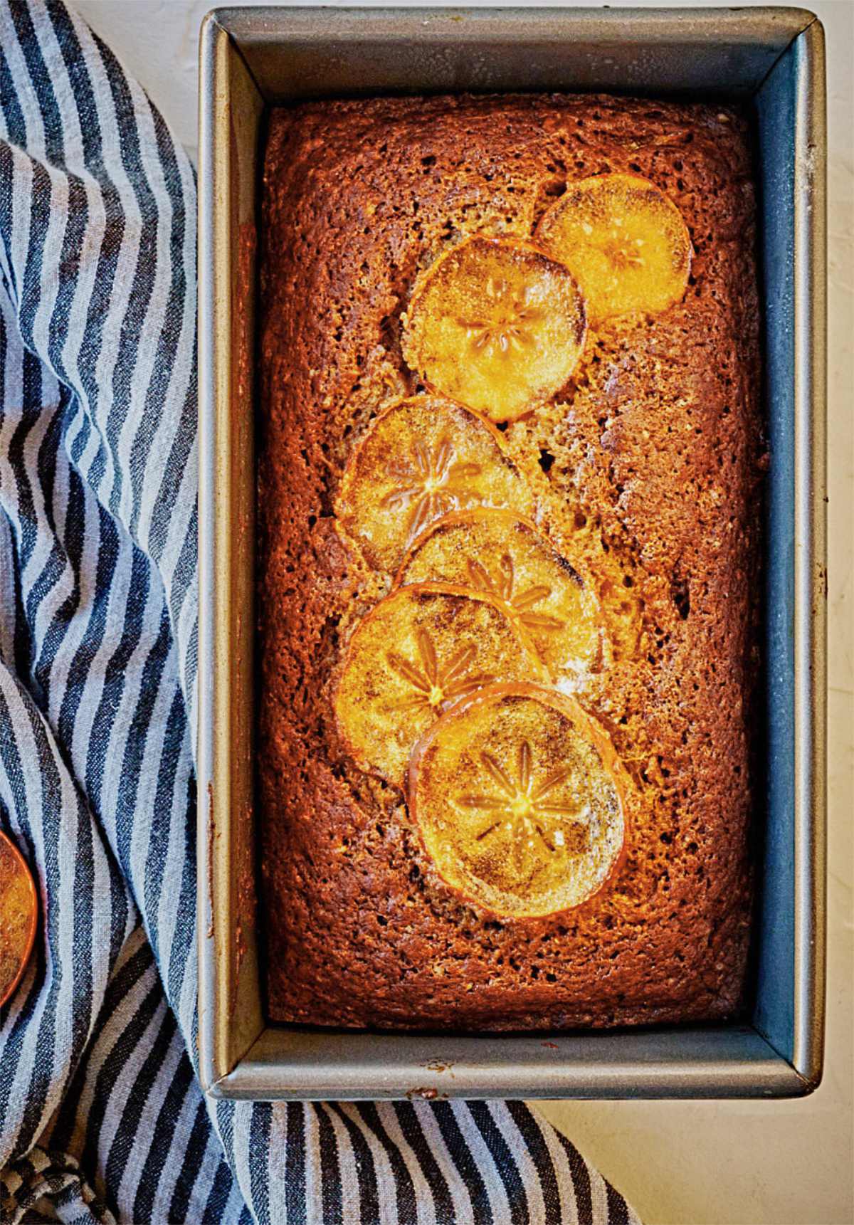 Baked quickbread loaf decorated with sliced persimmon next to a striped kitchen towel.