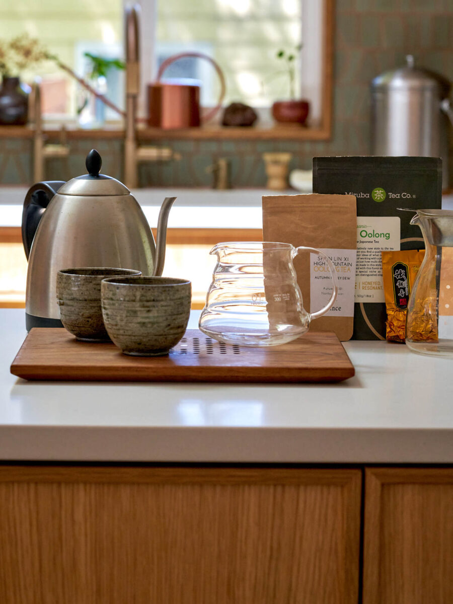 Tea kettle, cups, and glass pitchers set up on a countertop with different bags of tea behind them.